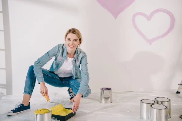 Belle jeune femme tenant rouleau de peinture et plateau à rouleaux avec de la peinture jaune tout en souriant à la caméra — Photo de stock