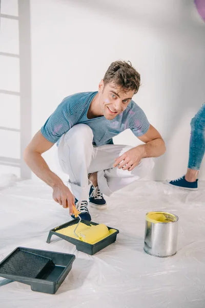 Beau jeune homme mettant rouleau de peinture dans le plateau de rouleau avec de la peinture jaune et souriant à la caméra — Photo de stock