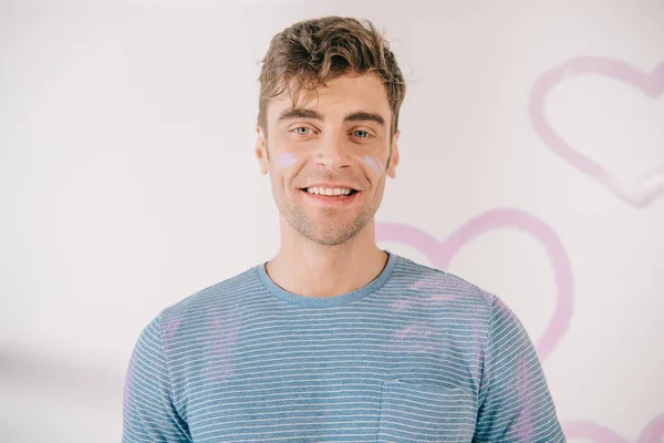 Beau jeune homme avec des taches de peinture rose sur le visage en regardant la caméra — Photo de stock