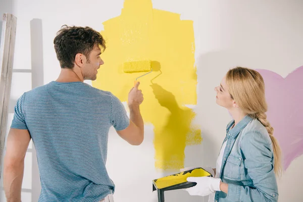 Jovem mulher segurando bandeja de rolo enquanto namorado pintura parede em amarelo com rolo de pintura — Fotografia de Stock