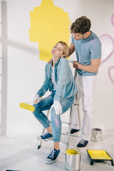 Handsome man standing near tired girlfriend sitting on ladder and holding yellow paint roller — Stock Photo