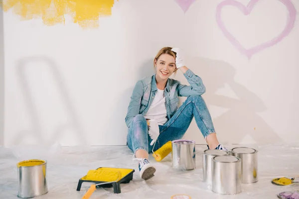 Atractiva mujer joven sosteniendo rodillo de pintura amarillo mientras está sentado en el suelo cerca de la pared - foto de stock