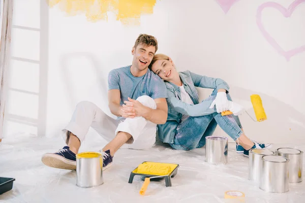 Happy young woman holding yellow paint roller while sitting on floor near boyfriend — Stock Photo