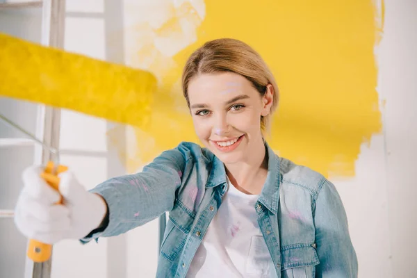 Вибірковий фокус красивої молодої жінки тримає жовтий валик для фарби і посміхається на камеру — стокове фото