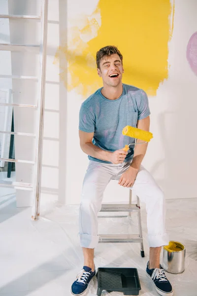 Schöner junger Mann sitzt auf Leiter, hält gelbe Farbrolle in der Hand und lächelt in die Kamera — Stockfoto