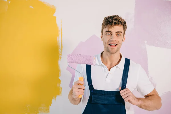 Handsome young painter in overalls holding paint roller while standing near wall painted in yellow and pink — Stock Photo