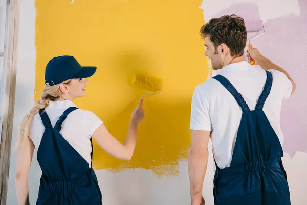 Pintores jóvenes en uniforme mirándose mientras pintan la pared en amarillo y rosa - foto de stock