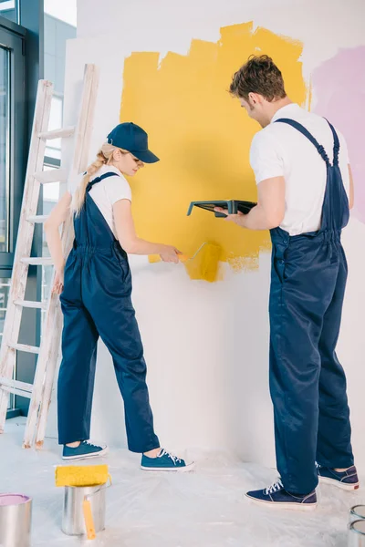 Joven pintor sosteniendo bandeja de rodillo mientras atractiva pared de pintura colega con rodillo de pintura - foto de stock