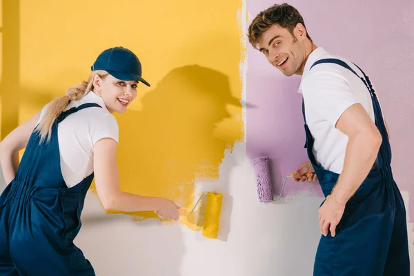 Два веселых художника смотрят в камеру, рисуя стену в желтом и розовом цвете — стоковое фото