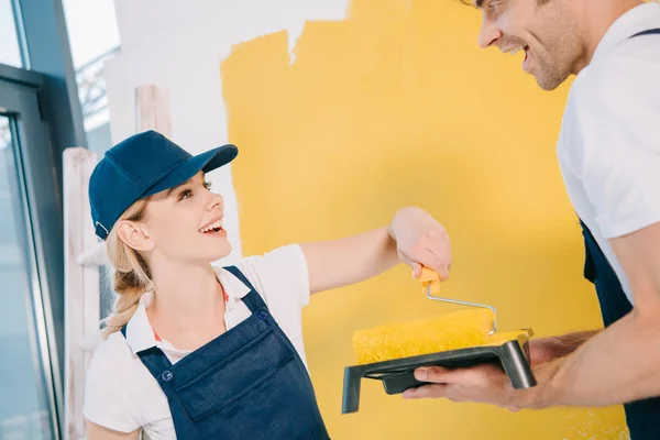 Alegre pintor en uniforme tomando la pintura de la bandeja de rodillo en las manos sobre el colega guapo - foto de stock