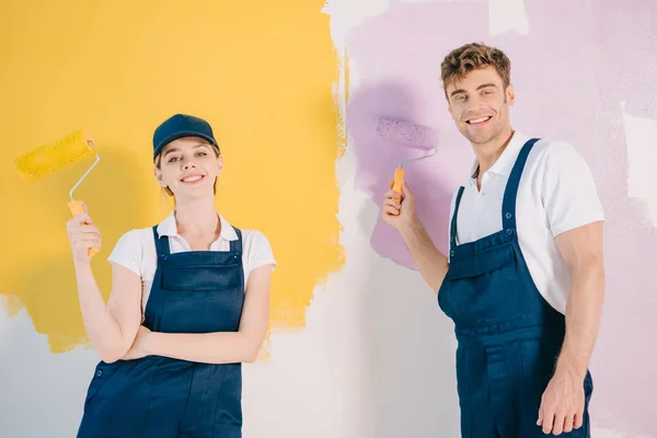 Jóvenes pintores con monos sosteniendo rodillos de pintura y sonriendo a la cámara - foto de stock