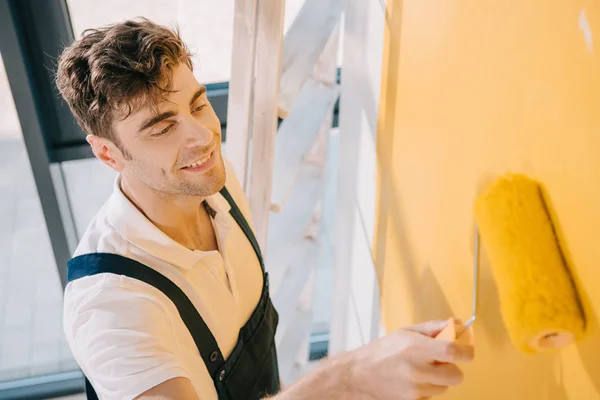 Joven pintor guapo sonriendo mientras pinta la pared en color amarillo con rodillo de pintura - foto de stock