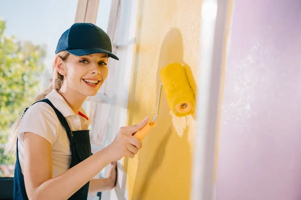 Allegro giovane pittore in uniforme sorridente alla macchina fotografica mentre dipinge parete in giallo e rosa con rullo di vernice — Foto stock