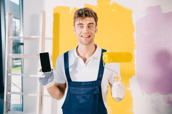 Alegre pintor en uniforme sosteniendo teléfono inteligente con pantalla en blanco y rodillo de pintura mientras sonríe a la cámara - foto de stock