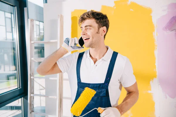 Joven pintor guapo hablando en el teléfono inteligente mientras sostiene el rodillo de pintura - foto de stock