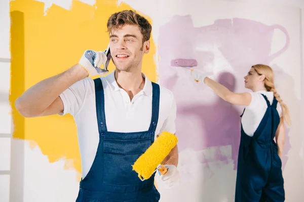 Pintor guapo hablando en el teléfono inteligente mientras atractivo colega pintura pared - foto de stock