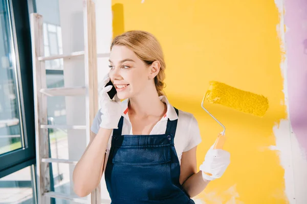 Atractivo joven pintor en uniforme hablando en el teléfono inteligente mientras sostiene el rodillo de pintura - foto de stock