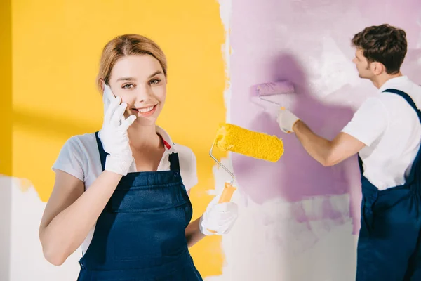 Pintor atraente em uniforme falando no smartphone enquanto bonito colega parede de pintura — Fotografia de Stock