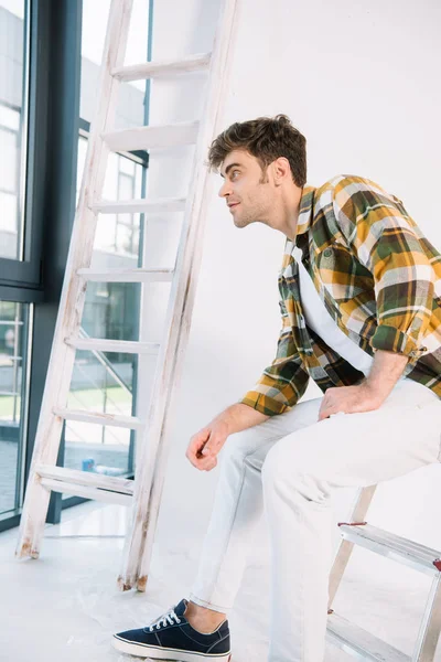 Schöner junger Mann auf Leiter sitzend und zu Hause wegschauend — Stockfoto