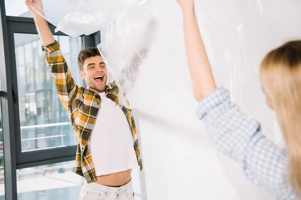 Feliz joven sosteniendo celofán con su novia mientras se prepara para la reparación de la habitación - foto de stock