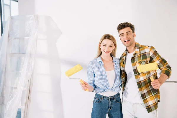 Feliz joven pareja sonriendo a la cámara mientras sostiene los rodillos de pintura amarilla - foto de stock