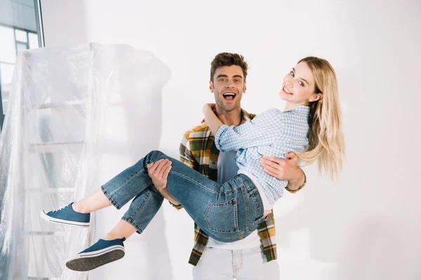 Happy young man holding girlfriend on hands while standing near white wall — Stock Photo