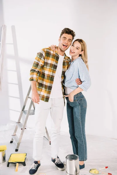 Happy young couple embracing and smiling at camera while standing near white wall — Stock Photo
