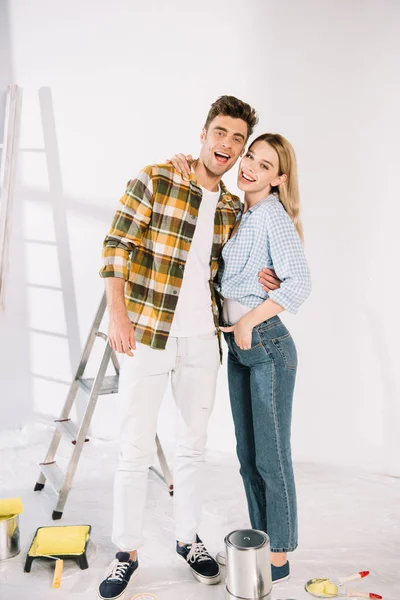 Feliz jovem casal abraçando e sorrindo para a câmera enquanto está perto da parede branca — Fotografia de Stock