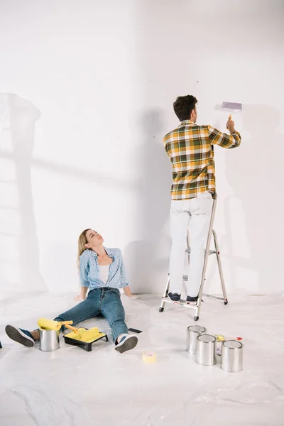 Mujer joven sentada en el suelo mientras su novio de pie en la escalera con rodillo de pintura rosa - foto de stock