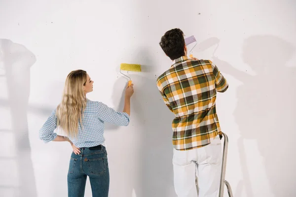 Mujer joven sosteniendo rodillo de pintura amarillo y novio sosteniendo rodillo de pintura rosa blanco de pie cerca de la pared blanca - foto de stock