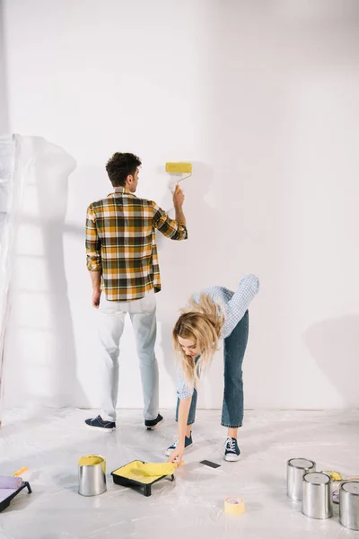 Jovem mulher colocando rolo de tinta na bandeja de rolo com tinta amarela, enquanto namorado pintura parede — Fotografia de Stock