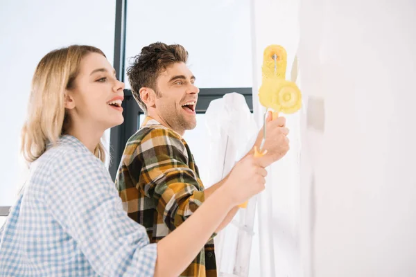 Feliz jovem casal pintura parede branca em amarelo com rolos de pintura — Fotografia de Stock