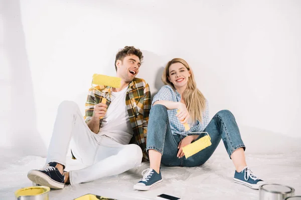 Heureux jeune couple assis près du mur blanc et tenant des rouleaux de peinture jaune — Photo de stock