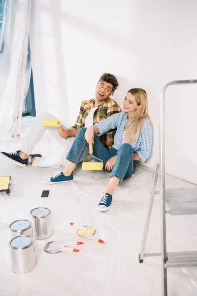 Cheerful young couple sitting near white wall and holding yellow paint rollers — Stock Photo