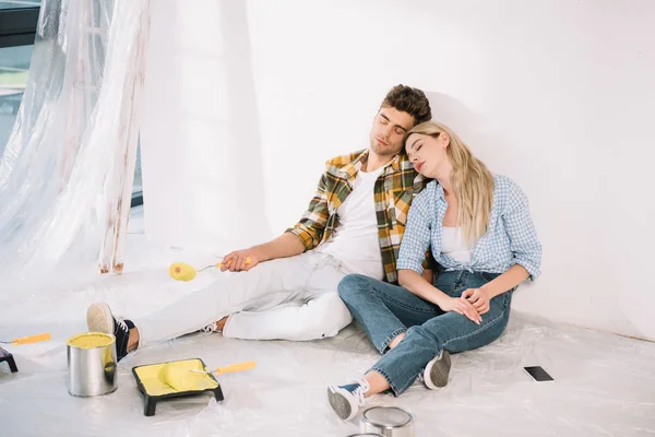 Couple épuisé dormant assis par un mur blanc près d'un plateau à rouleaux avec de la peinture jaune — Photo de stock