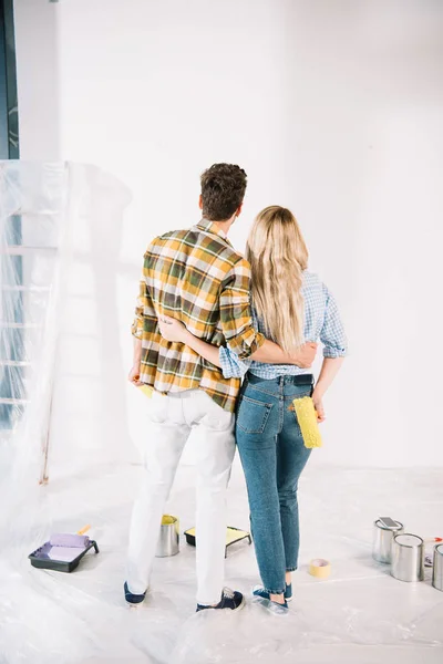 Rückansicht eines jungen Paares, das sich umarmt, während es die weiße Wand zu Hause betrachtet — Stockfoto