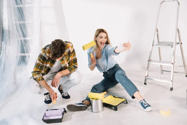 Cheerful young woman holding yellow paint roller and taking selfie while boyfriend putting paint roller into pink paint — Stock Photo