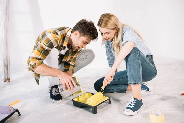 Joven agregando pintura amarilla en la bandeja de rodillos mientras su novia sostiene el rodillo de pintura - foto de stock