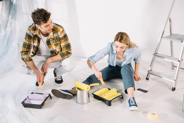 Handsome man holding pink paint roller and woman putting paint roller into yellow paint while sitting on floor — Stock Photo