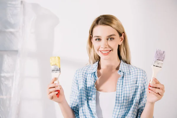 Attractive young woman holding yellow and pink paintbrushes and smiling at camera — Stock Photo