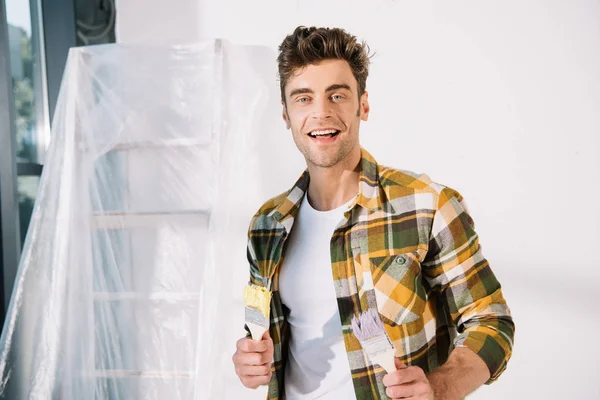 Cheerful young man looking at camera while holding yellow and pink paintbrushes — Stock Photo