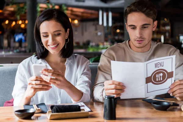 Femme souriant tout en utilisant smartphone près de l'homme avec menu — Photo de stock