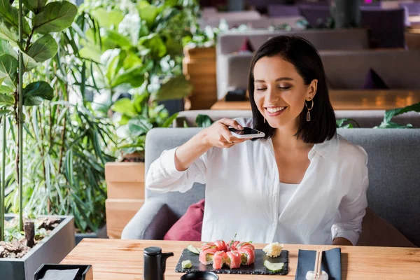 Femme heureuse prenant des photos de sushi savoureux au restaurant — Photo de stock