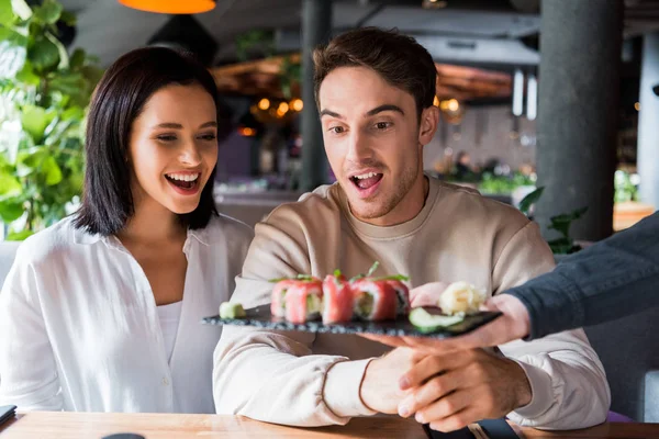 Vista ritagliata di cameriere che tiene il piatto con sushi saporito vicino a coppia sorpresa in sushi bar — Foto stock