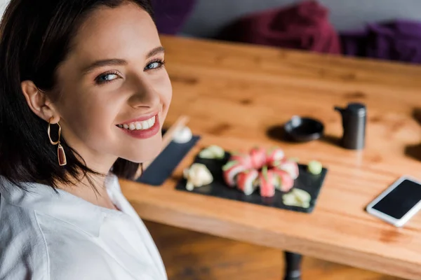 Избирательный фокус счастливой женщины возле смартфона с чистым экраном и вкусными суши в ресторане — стоковое фото