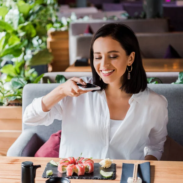 Happy woman taking photo of delicious sushi in restaurant — Stock Photo