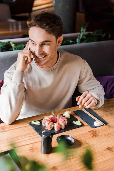 Enfoque selectivo de hombre feliz hablando en el teléfono inteligente en el bar de sushi cerca de la comida sabrosa - foto de stock