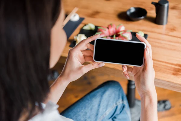 Enfoque selectivo de la mujer sosteniendo teléfono inteligente con pantalla en blanco cerca de sushi en el restaurante - foto de stock