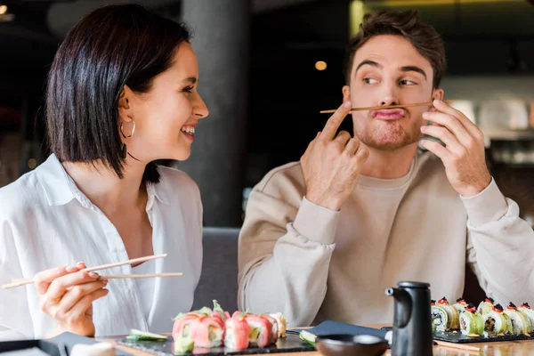 Foyer sélectif de l'homme tenant baguettes près du visage tout en étant assis avec fille gaie dans le bar à sushi — Photo de stock