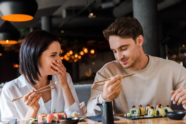 Uomo felice che tiene le bacchette vicino alla donna allegra che ride mentre copre il viso nel ristorante — Foto stock